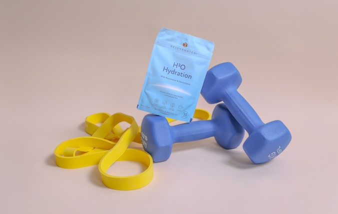 A pouch of Rejuvenated's H3O Hydration product next to a pair of blue dumbbells and a yellow resistance band.